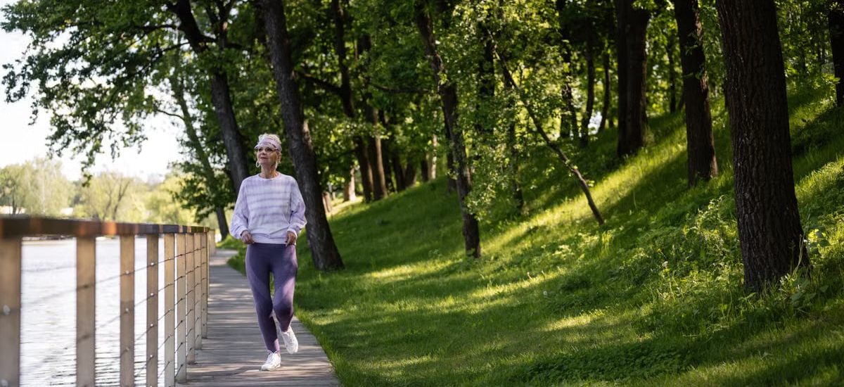 Elderly woman running along a riverside path in a green park, embracing fitness and nature.