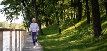 Elderly woman running along a riverside path in a green park, embracing fitness and nature.