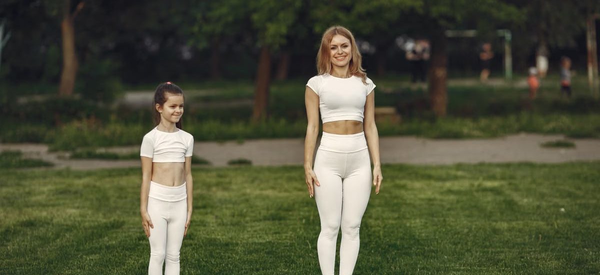 A woman and girl in white activewear standing on a grass field outdoors, smiling.